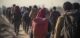 Group of refugees standing in a barren landscape, wearing backpacks. Harsh lighting casts long shadows, symbolizing displacement and crisis. A diverse and resilient community, united in their journey